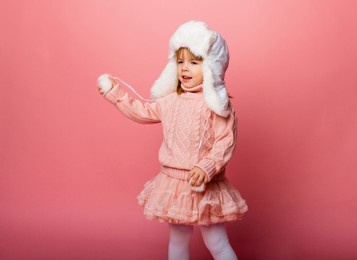 little blonde girl in winter clothes and a fur hat on a pink background.