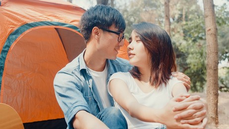 young asia campers couple sitting in chairs by tent in forest. male and female traveler relaxing and talking on a summer day at campsite. outdoor activity, adventure travel, or holiday vacation.