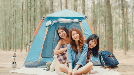 group of young asia camper friends camping near relax enjoy moment in forest. female traveler using smartphone taking selfie at campsite. outdoor activity, adventure travel, or holiday vacation.