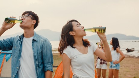 group of asia best friends teenagers focus on couple toast beer enjoy camping party with happy moments together beside tents in national park. on the background beautiful nature, mountains and lake.