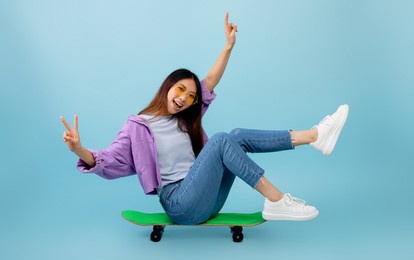 joyful asian lady sitting on skateboard with legs up and smiling to camera, showing v-sign gesture. young woman spending time and having fun over blue studio background