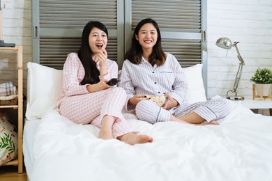 pajama party entertainment and junk food concept. two happy asian japanese women in sleepwear sitting on white bed and enjoy comedy laughing together. relax female eating bowl of popcorn and watch tv