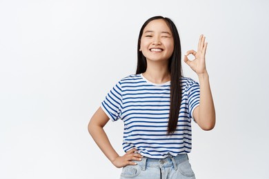 image of teen asian girl winking and smiling, showing okay, ok gesture, assuring you, confirm something is good, confident in quality, standing over white background