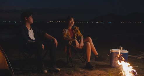 young asia couple backpackers enjoying in camping at night near campfire on beach. male and female traveler relaxing and talking at campsite. outdoor activity, adventure travel, or holiday vacation.