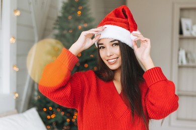 young beautiful asian woman with dark long hair in cozy red knitted sweater and santa hat having fun in room with christmas tree