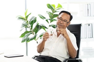 senior asian man with cup of water and pill taking remedy in cozy room at home. old male taking medicine