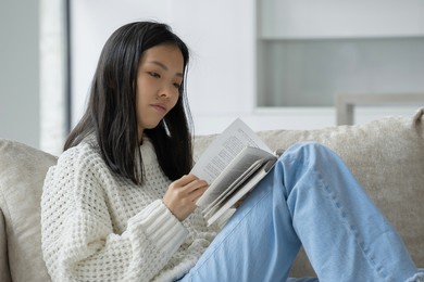 happy asian woman in a white sweater sitting on the couch and reading a book at home