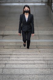 a thai asian woman wearing a black suit, trousers and a mask. she was walking up the stairs. in the concept of travel and advancement of a business woman or office worker.