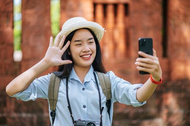 young asian backpacker female wearing hat traveling in historic site, she use smartphone and camera take a photo with happy