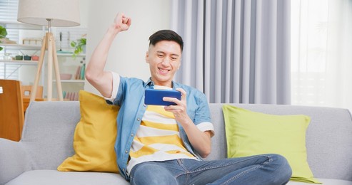 asian young man sitting on sofa playing game by mobile phone in living room at home - fist gesture