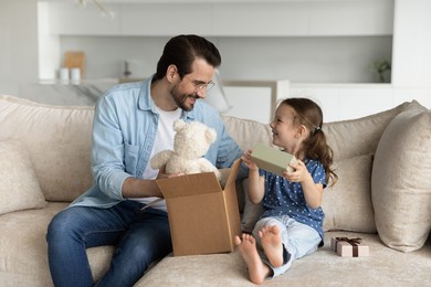 happy young father unboxing parcel with gifts for small cute kid daughter, sitting together on cozy sofa in living room, satisfied with fast delivery purchase shipment or high quality fluffy toy.