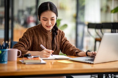 young attractive university student using a laptop computer, studying online at home. cheerful caucasian asian woman writes notes, planning working process, sitting at home. exam preparation.