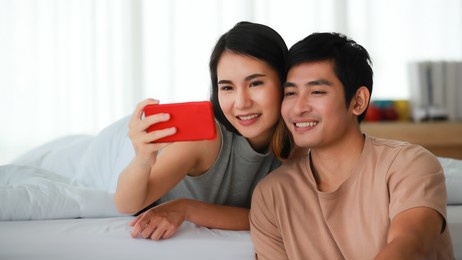 portrait shot of asian lover couple relaxes at home on the holidays. the wife lies on bed behind her husband that sits on the floor and embracing him. woman playing social media on a smartphone