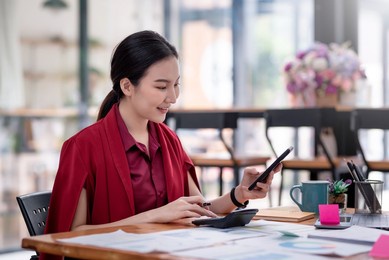 side view of young asian businesswoman holding a smartphone using a calculator to calculate work graph papers placed on the table.