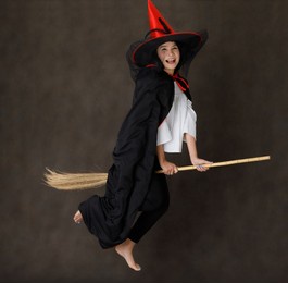 full body of happy young asian female in witch costume with bare feet pretending to fly on broom during halloween party against dark gray background