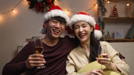 happy young sweet couple in santa hat making video call and talking to camera greeting family on christmas and new year holidays. two female and male lovers sitting on couch with glasses of wine