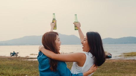 group of asia girl couple best friends teenagers drinking have fun salute toast of bottle beer enjoy party with happy moments together in campsite. on background beautiful nature, mountains and lake.