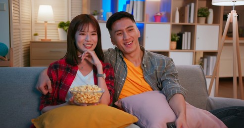 asian young couple eating pop corn are watching tv and sitting on the couch happily in night living room at home