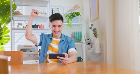 asian young man sitting on sofa playing video game by mobile phone in dining room at home - fist gesture