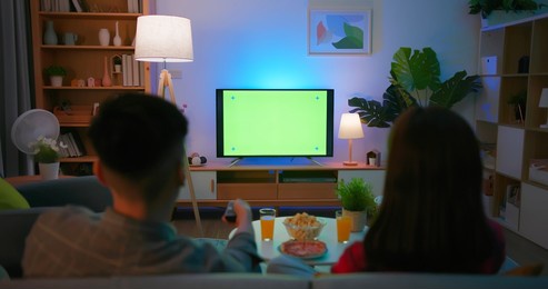 back view of asian young couple are watching green chroma key screen tv with food and drinks fun while sitting on couch at night in the living room