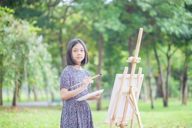 asian kid painting on the canvas in the park. happy child girl drawing a picture outdoors