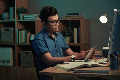 serious young vietnamese programmer in eyeglasses using digital tablet in dark home office