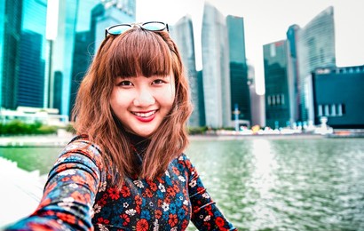 young asian woman taking happy selfie traveling at singapore skyline - wanderlust life style concept with millenial girl having fun by urban city surrounds - vivid bright filter with focus on face