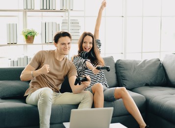 playful couple hold gamepad controller playing video games as home entertainment. joyful excited two friends sit on sofa in living room having fun with videogame indoor hobby leisure activity together
