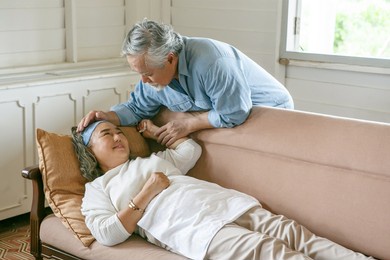 happy old asian husband take care and embracing elderly wife suffering pain of sick lying down on sofa in living room at home . lovely asia senior couple in love support together on couch 