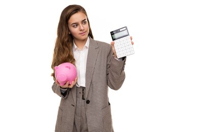 photo of beautiful cute pretty asking thoughtful brunette teenage girl with long hair in stylish plaid jacket, plaid pants and white shirt holds calculator and pink pig piggy bank isolated on white