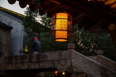 A lighted lantern hanging under the eave with texts which means "Lili ancient town"; meanwhile a man wearing a pink hat walks across the stone bridge in the back.