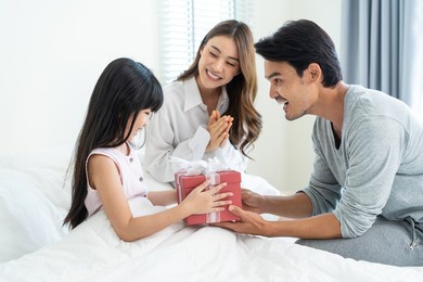 asian happy family, young parents surprise sleep daughter with gift. attractive man and woman couple wake up their little kid from bed in bedroom to give birthday present and celebrate tradition party