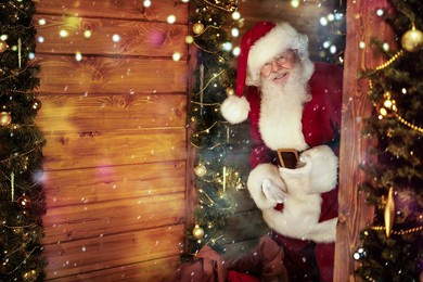 cheerful santa claus looks into a wooden house, beautifully decorated for christmas, with a big bag of presents. 