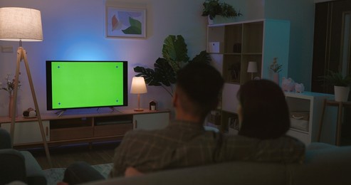 rear view of asian young couple are watching green chroma key screen tv and having fun while sitting on couch at night in the living room