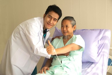 happy asian male patient lies on the bed, and raises her thumb up. when a doctor uses a stethoscope to listen to the lungs. concept of believe in treatment.
