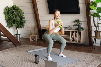 fit and healthy asian woman doing squats at home, workout fitness training session with dumbbells and protein shaker in living room, watching sports video on laptop
