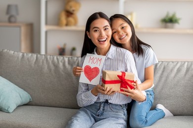 mother's day gift. happy asian girl greeting young surprised mom, giving her handmade card and wrapped gift box, sitting on sofa at home interior and smiling at camera