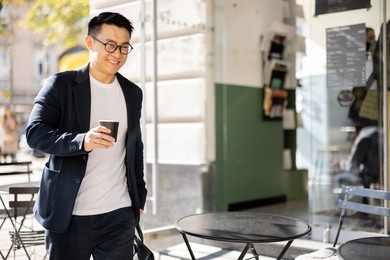 asian businessman with coffee going from entrance of building. smiling modern adult successful man wearing suit and glasses with briefcase. city at sunny day