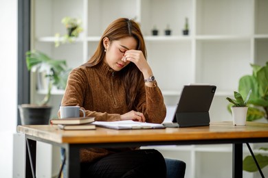 asian women sitting in a home office with stress and eye strain.tired businesswoman holding eyeglasses and massaging nose bridge. there are tablets, laptops, and coffee.