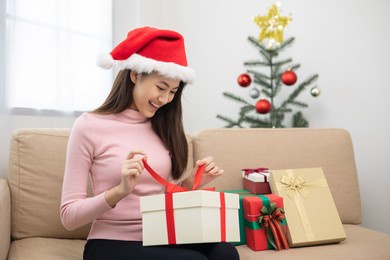 beautiful asian woman unpack the present gift box pull ribbon guess what inside the box. young happy cute girl celebrate in christmas holiday festival with many gift box sitting on sofa at home
