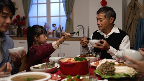 happy asian grandfather talking with hand gestures as his polite granddaughter putting food into his bowl at dinner table on chinese new year's eve at home