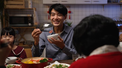 selective focus smiling asian father talking to family while enjoying tasty food at dinner table. people chatting happily during reunion meal on lunar new year's eve at home