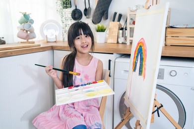 portrait of asian young kid coloring and painting board in living room. little adorable child learn how to draw art picture with watercolor and brush on holiday at home and smile, looking at camera.