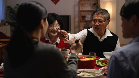 selective focus with over shoulder view smiling asian elderly couple grandfather and grandmother holding food with chopsticks and giving to their daughter in law at dinner table on eve of new year