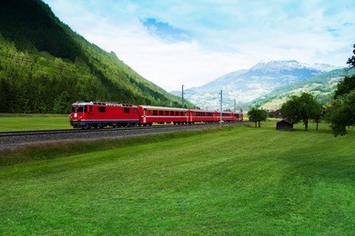 long red train crossing plain green valley near alps, switzerland in summer