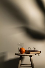 still life of orange fruit, glass of water, a book and glasses on white wooden stool against white wall with sunlight and shadow