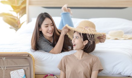 asian teenager traveler girl sitting smiling on carpet floor and young friend lying down on bed booking reserve hotel online with laptop notebook computer together in bedroom at home before travel.