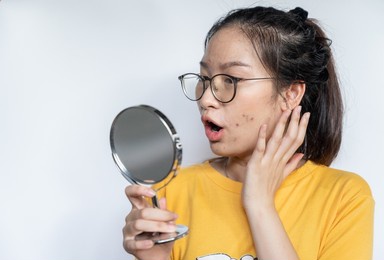 young asian woman feeling shock while looking acne and scar occur on face by mini mirror. conceptual shot of acne and problem skin on female face.
