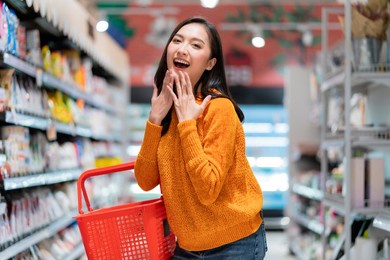 smiling cheerful and joyful female woman hand hold shopping basket hand gesture greeting surprise standing between supermarket product shelf aisel convenience store supermarket department store mall
