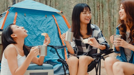 group of young asia camper friends sitting in chairs by tent in forest. teenager girl traveler relax and talk on a summer day at campsite. outdoor activity, adventure travel or holiday vacation.